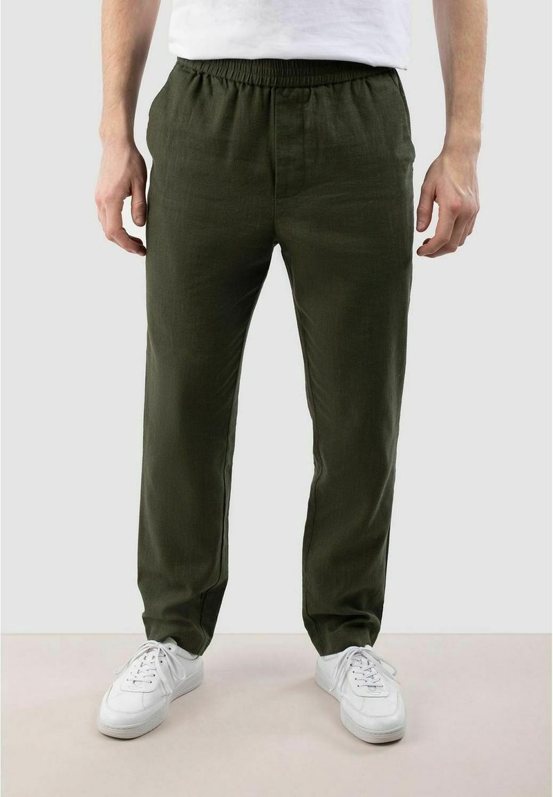Homme portant un pantalon vert olive à taille élastique et des baskets blanches, debout devant un fond clair uni.