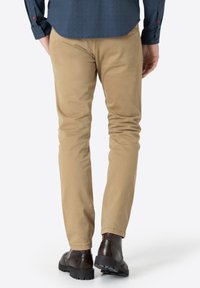 Mann, der beigen Chinos, dunkelbraune Leder-Brogue-Stiefel und ein navyblaues Hemd mit roten und blauen Knöpfen trägt, von hinten gezeigt.