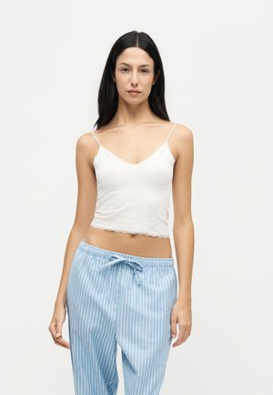 Femme aux longs cheveux noirs portant un débardeur court blanc et un pantalon bleu clair rayé avec cordon, debout devant un fond uni.