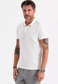 Vit pikétröja med klassisk krage och korta ärmar, tillverkad av mjuk bomull. Utmärkande tatueringar på armarna och ett silverur på handleden.