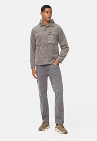 Veste en velours côtelé gris avec capuche, fermeture à boutons sur le devant, deux poches poitrine et une poche contrastante verte. Portée avec un pantalon gris et des baskets.