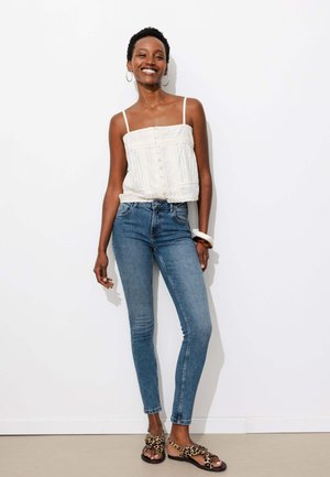 Femme souriante portant un haut blanc sans manches, un jean skinny bleu, des sandales imprimé léopard et des boucles d’oreilles créoles, debout contre un mur blanc.
