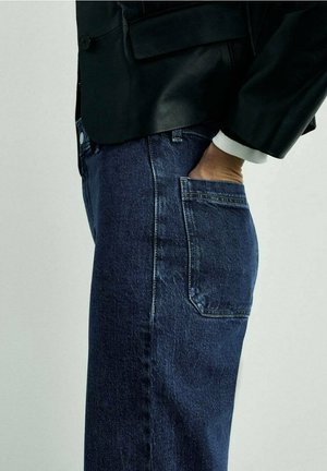 Donkere denim jeans met een rechte pasvorm, voorzien van een achterzak en subtiele vervaging. Gecombineerd met een zwarte leren blazer en witte manchetten.