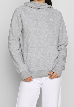 Osoba ubrana w jasnoszarą bluzę Nike z kapturem i kieszenią z przodu oraz białe spodnie, stojąca na jednolitym tle.