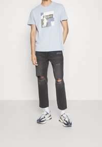 Camiseta de manga corta azul claro con un estampado gráfico. Llevada con jeans negros desgastados y zapatillas con acentos en azul marino.