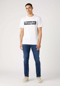 Camiseta blanca con un logo rectangular "Wrangler" en negro, combinada con jeans azules y zapatos beige. Modelo de pie frente a un fondo claro.