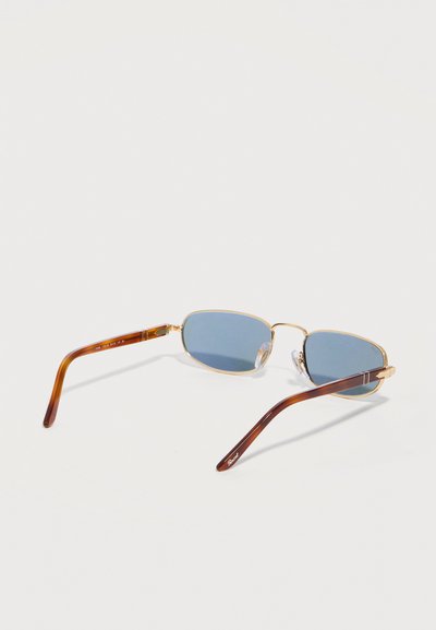 Γυαλιά ηλίου με χρυσά ορθογώνια πλαίσια, μπλε φακούς και καφέ βραχίονες σε σχέδιο χελώνας που ακουμπούν σε λευκή επιφάνεια.