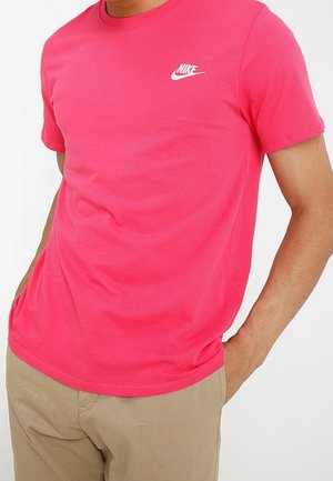 Mann trägt ein leuchtend pinkes Nike-T-Shirt mit beigen Hosen, die Hände lässig in den Taschen, vor einem schlichten weißen Hintergrund.