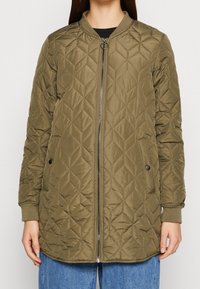 Lang, olivengrøn quiltet jakke med frontlynlås, ribbet krave og manchetter samt sidelommer med trykknapper, båret over blå jeans.