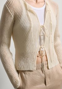Helle beige Strickjacke mit vertikalen Rippen, langen Ärmeln und einer Bindeschließe vorne. Über einem weißen, figurbetonten Oberteil getragen.