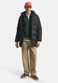 Zwarte pufferjas met een capuchon, groene trui eronder, beige broek en bruine schoenen. De man draagt een oranje pet.