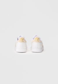 Sapatilhas de couro branco com detalhes em camurça bege suave, apresentando um logótipo dourado no calcanhar. Design limpo, sola plana de borracha branca.