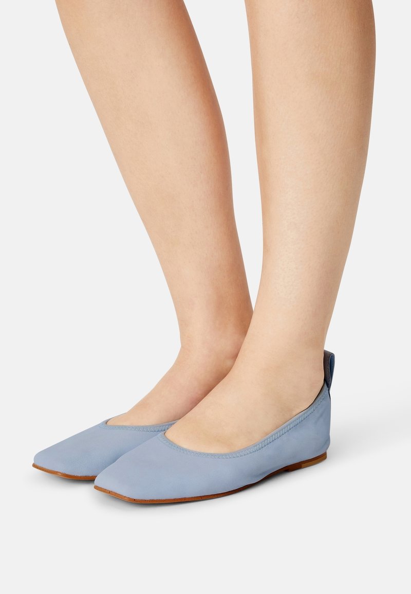 Jambes portant des chaussures plates bleu clair à bout pointu avec des semelles marron sur fond blanc.