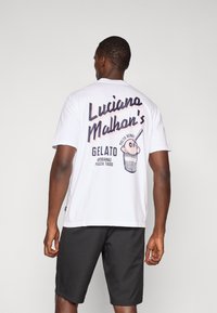 White cotton t-shirt with large black text reading "Luciano Malhan's" and an illustrated ice cream cone on the back. Paired with black shorts.