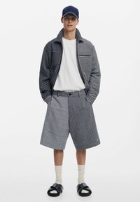 Csíkos kék és fehér zakó és rövidnadrág, fehér pólóval és fekete szandállal párosítva. Az öltözet laza illeszkedésű, és zsebekkel rendelkezik.