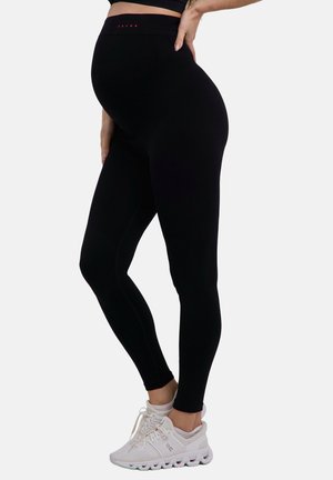 Donna incinta che indossa leggings premaman neri e scarpe da ginnastica bianche, in piedi con una mano sul fianco davanti a uno sfondo bianco.