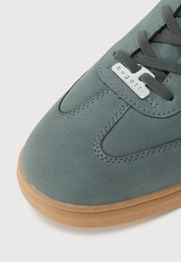 Zapatillas de ante gris con punta redonda, que cuentan con una suela de goma, costuras de contraste y una etiqueta de logo metálica en el lateral.