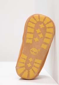 Brązowa podeszwa zamszowego buta z żółtą gumową podeszwą zewnętrzną ozdobioną wzorem krzyża i pasków. Gładka tekstura z wzmocnionymi krawędziami.