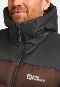 Uomo che indossa una giacca imbottita Jack Wolfskin marrone e nera con un alto colletto chiuso con cerniera.