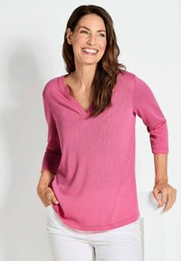 Helles rosa Langarmshirt mit V-Ausschnitt, weichem Material, lässiger Passform und abgerundetem Saum, kombiniert mit weißen Hosen.