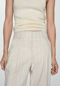 Pantalons rayés beige avec une coupe ajustée, comportant des plis sur le devant, des poches latérales et une texture de tissu légère.