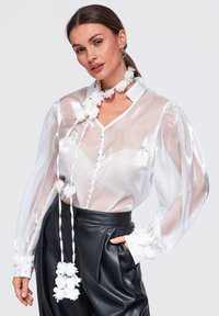 Femme portant un chemisier blanc transparent à manches longues avec une écharpe fleurie et un pantalon en cuir noir taille haute, posant avec une main dans la poche.