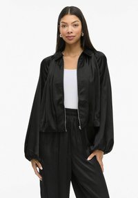 Černá saténová bomber bunda s krátkým střihem, zapínáním na zip vpředu a balónovými rukávy. Stylizováno přes bílý top s volnějším střihem.