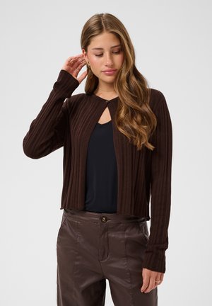 Jeune femme aux longs cheveux ondulés portant un cardigan côtelé marron foncé, un haut noir et un pantalon en cuir marron, touchant ses cheveux et regardant vers le bas.