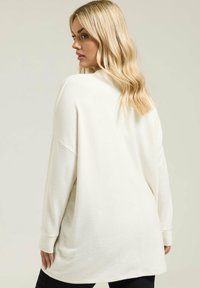 Blondynka w oversize'owym kremowym swetrze, odwrócona plecami, spogląda na bok na tle jasnego jednolitego tła.