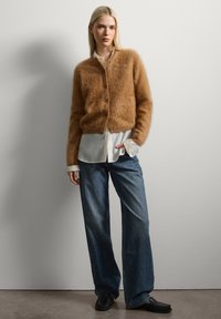Brauner, flauschiger Cardigan mit Knopfleiste, über einem weißen Hemd getragen. Weite blaue Jeans mit weitem Bein, kombiniert mit schwarzen Slippern.