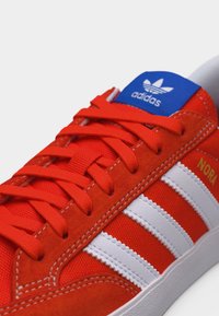 Κόκκινο αθλητικό παπούτσι Adidas με λευκές ρίγες, μπλε ετικέτα στη γλώσσα, κόκκινα κορδόνια και τη λέξη «NORA» τυπωμένη σε χρυσό στη πλευρά.