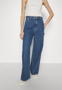 Jeans in denim blu a gamba larga con vita alta, design classico a cinque tasche e cuciture a contrasto. Indossati con un top bianco cropped e scarpe chiare.