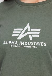 Grøn bomulds t-shirt med et hvidt logo med striber og teksten "ALPHA INDUSTRIES KNOXVILLE, TENNESSEE, U.S.A." på forsiden.