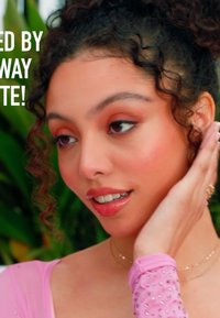 Close-up van een persoon met krullend haar, gekleed in een doorschijnende roze top versierd met rhinestones. Zachte make-up met warme tinten en glanzende lippen.