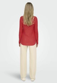 Maglione rosso a maniche lunghe con design a maglia testurizzata, abbinato a pantaloni bianchi a gamba larga. Il maglione presenta un orlo curvo e maniche aderenti.