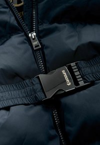 Tumši zils izolēts jaka ar centrālu rāvējslēdzēju un melnu plastmasas sprādzi ar "Superdry" logotipu. Strukturēta auduma ar quilted dizainu.