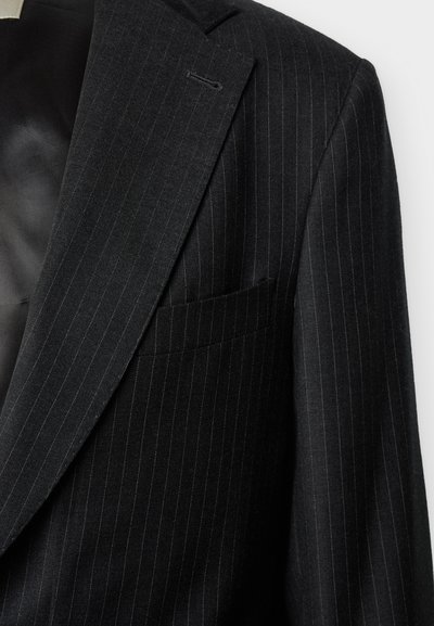 Črno suknjič s črtami, z izrezanim reverjem in prsno žepom, prikazan delno spredaj in ramo na belem ozadju.