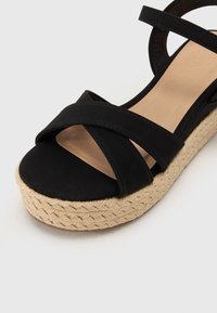 Crne espadrille sandale s ukriženim trakama, teksturiranom jute platformom i glatkom bež uloškom.