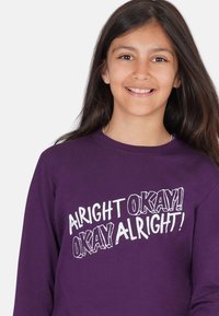 Lila Langarmshirt mit weißem Grafiktext: "ALRIGHT OKAY! OKAY ALRIGHT!" in verschiedenen fetten Stilen. Glatte Baumwolltextur.