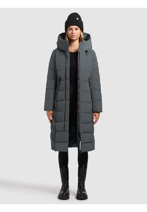 Frau in einem langen grauen gepolsterten Mantel mit Kapuze, schwarzem Beanie, schwarzen Leggings und schwarzen Stiefeln, die vor einem schlichten Hintergrund steht.