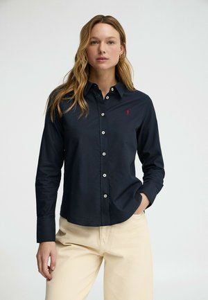 Mujer vestida con una camisa azul marino con botones y un logo rojo en el pecho, y pantalones beige, de pie con una mano en el bolsillo contra un fondo liso.