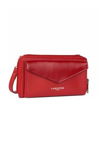 Portefeuille en cuir rouge avec une finition texturée, comportant un rabat triangulaire, une fermeture zippée et une bandoulière. Le nom de la marque "LANCASTER PARIS" est affiché.