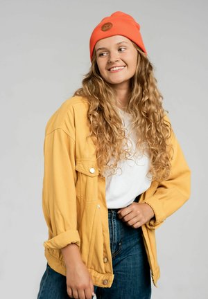 Żółta, oversize'owa dżinsowa kurtka z srebrnymi guzikami, biała koszulka, niebieskie dżinsy oraz pomarańczowa czapka beanie z brązową naszywką.