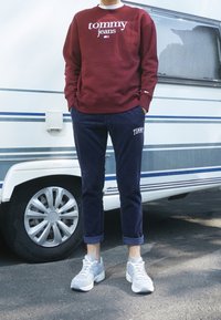 Felpa bordeaux con logo "tommy jeans", pantaloni blu navy con ricamo "TOMMY" e sneaker grigie. Outfit casual con uno sfondo di un camper.
