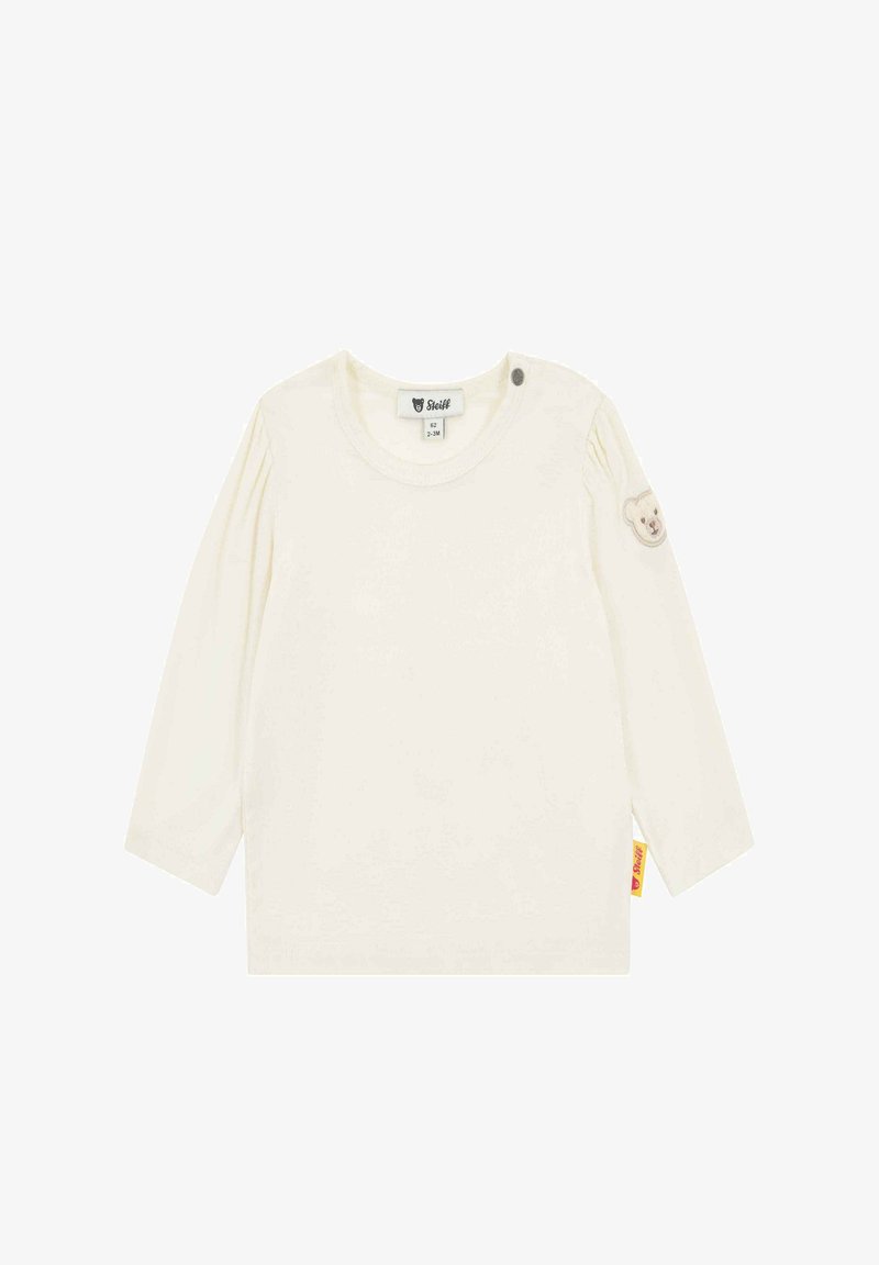 Chemise à manches longues crème en coton doux, avec un col rond, un détail de boutons sur l'épaule et un patch ours sur la manche.
