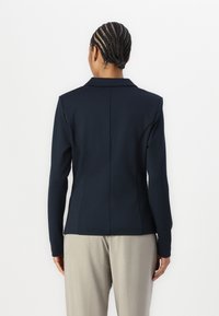 Blazer ajusté bleu marine avec un col structuré, des manches longues et une texture lisse. Détails de coutures remarquables au dos pour améliorer la forme.