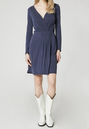 Frau trägt ein knielanges, marineblaues Wickelkleid mit langen Ärmeln und weißen Cowboy-Stiefeln bis zur Mitte der Wade und steht vor einem schlichten Hintergrund.