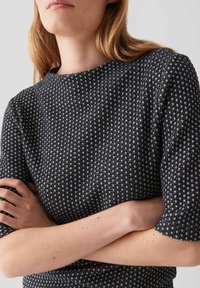 Femme à la peau claire et aux cheveux blonds portant un pull en maille noir et blanc à manches courtes, les bras croisés sur le ventre.