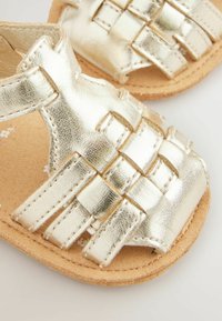 Gold metallic woven sandals with a flat, textured cork sole. Features a secure strap design and decorative stitching accents.