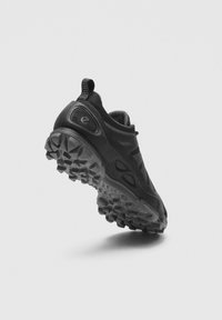 Juoda bėgimo lauko batelio pusė, rodanti tvirtą pado raštą ir tekstūruotą viršutinę dalį su kilpelėle ant šviesiai pilkos fono.